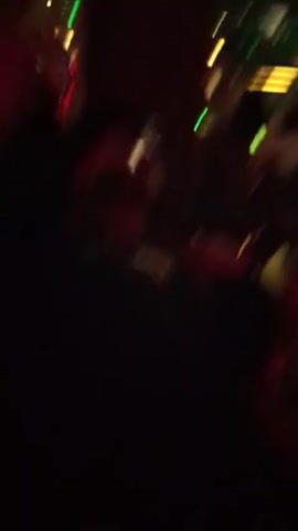The video shows two men in a dark bar or club. The camera is held by one man, who is moving around and singing along to music. He is wearing a dark shirt. The other man is standing next to him, also wearing a dark shirt. He looks at the camera and smiles. The environment is dimly lit with red and green lights, and there are other people in the background. There are light fixtures visible on the ceiling.
