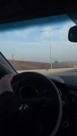 The video shows a car's dashboard view while driving on a highway. The driver's left hand is visible on the steering wheel. On the dashboard, there is a phone holder and a hanging ornament. Outside, the landscape consists of a brown, hilly terrain and a pale blue sky with some clouds. There are street lights lining the highway, and other vehicles, including large trucks, are on the road. The car is a Hyundai, as indicated by the logo on the steering wheel.