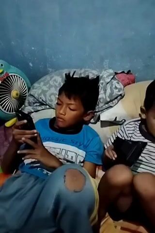 A boy in a blue t-shirt is playing on a smartphone. His knees are ripped on his pants. Another boy is sitting next to him playing a handheld video game. They are both sitting on a bed in a dimly lit room.