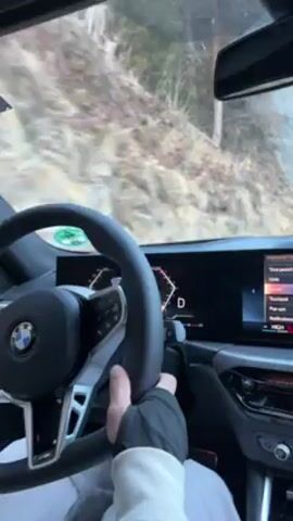 The video shows a person driving a BMW on a snow-covered road. The view is from inside the car, focused on the steering wheel, dashboard, and the road ahead. The person is wearing a white sweatshirt and a black fingerless glove on their left hand. The dashboard displays digital information including speed. Trees are visible along the side of the road. The car is moving slowly on the partially snow-covered asphalt.