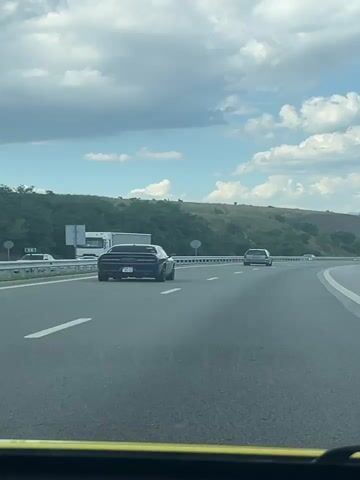 This media file captures a scenic drive on a highway, likely in Blatino, Bulgaria, under a bright, cloudy sky. The perspective is from inside a vehicle, looking forward.

The primary subjects are two cars on the road. In the foreground, a dark-colored, classic-style sports car with a distinctive angular design is moving away from the camera in the left lane. Its rear is clearly visible, including its taillights and license plate which appears to have text and numbers. Further ahead in the right lane, a lighter-colored sedan is also traveling in the same direction. A white semi-trailer truck is visible in the far left lane, moving in the opposite direction.

The setting is a multi-lane highway bordered by a metal guardrail on the left. Beyond the guardrail, rolling hills covered in lush green vegetation rise up, with some trees dotting the landscape. The road surface is asphalt, with clear white lane markings. The time of day appears to be daytime, given the bright sky and clear visibility. The weather seems pleasant, with fluffy white clouds scattered across a blue sky.

The overall mood conveyed is one of travel and freedom, with the open road and expansive sky contributing to a sense of journey. There are no discernible emotions expressed, as the focus is on the external environment and the vehicles.

Visible text includes what appears to be a license plate on the sports car and potentially signage on the truck and along the roadside, though these are not clearly legible. The yellow frame at the bottom suggests the media might be a phone recording.
