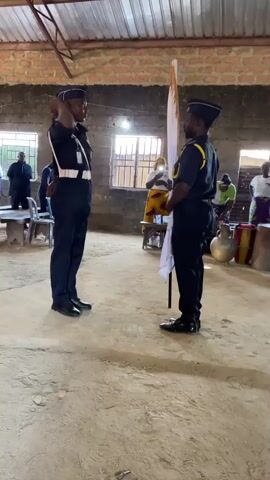 Two men in dark blue uniforms and caps stand facing each other in a room with a dirt floor and a corrugated metal roof. The man on the left faces the camera, while the man on the right faces away from the camera, holding a white flag with a light brown design on a pole. He is handing the flag to the man on the left. In the background, several other people are visible, some seated and some standing. The room appears to be a communal gathering space, possibly a hall or a church. The overall scene suggests a formal handover or presentation ceremony.