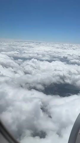 The video shows an aerial view from an airplane window, looking down at a vast expanse of fluffy white clouds. The sky above is a clear, bright blue. Patches of land with fields and bodies of water are visible through gaps in the clouds. The edge of the airplane's engine nacelle is visible in the lower right corner of the frame. The camera is mostly static, with slight movements consistent with being on an airplane in flight. The lighting suggests it is daytime, with the sun illuminating the clouds from above. The overall impression is one of peaceful flight above the weather.