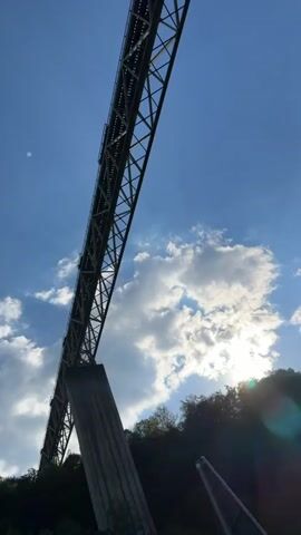 The video shows a long steel bridge with a blue sky and clouds as a backdrop. The bridge has a lattice structure, and one of its support columns is visible on the left side of the frame. The sun is shining behind the clouds, creating a lens flare effect. Trees are visible at the bottom of the frame, below the bridge. A road with a guardrail runs along the bottom of the frame, and a black car passes by on the road.