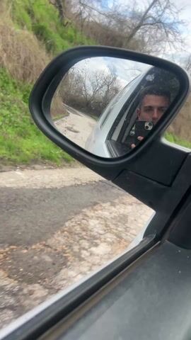 A first-person perspective video shows a person driving a car on a narrow road surrounded by green vegetation and bare trees. The road is paved but cracked and uneven. The driver is reflected in the side mirror, holding a smartphone. The video then transitions to the dashboard view, showing the driver's hands on the steering wheel. In the background, a mountain can be seen. The weather appears cloudy or overcast.
