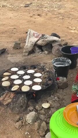 The video shows a woman cooking small pancakes on a griddle over an open fire. She is wearing a colorful headscarf and dress. There are several buckets and a plate with a spoon nearby. The ground is dirt and there are some rocks and a bag of something in the background. A chicken is visible in the background.