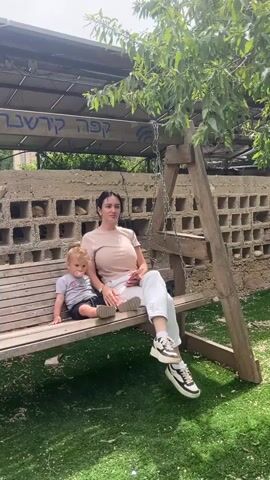 A woman and a young child are sitting on a wooden swing outside on a sunny day. The woman has dark hair pulled back, a light brown short-sleeved shirt, white pants, and black and white tennis shoes. The child is wearing a gray t-shirt and dark shorts. The swing is under a tree, and behind the swing is a wall made of cinderblocks. There is a covered area above the swing, with writing in Hebrew on it. The ground is covered in artificial turf.