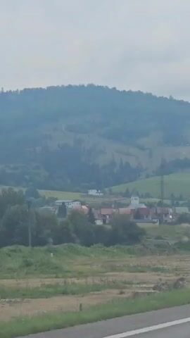 The video shows a view from a moving car, focusing on a rural landscape. In the foreground, the hood of a white car is visible, with a license plate that reads "CNA 1V77". The car appears to be driving on a paved road. Beyond the road, a field of dry grass and dirt is seen, followed by a small village nestled in a valley. The village consists of several houses with red roofs, some trees, and a white building that resembles a church. In the background, rolling hills covered in green trees rise up, and the sky is overcast with a pale, hazy light. The overall impression is of a scenic drive through the countryside on a cloudy day.