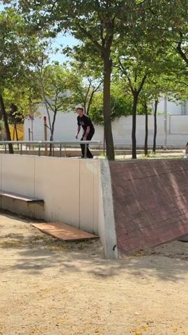 A skateboarder in a black t-shirt and black pants approaches a concrete ledge with a metal rail. They ollie onto the rail and grind across it, then ollie off the end of the ledge and onto a sloped brick surface. The skateboarder dismounts the skateboard, and it lands on the ground. The skateboarder walks away from the skateboard. In the background, there are trees, a white wall, and people sitting and watching. The ground is covered in sand and dirt.