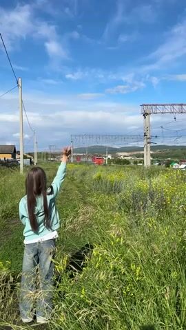 A young person stands in a field of tall green grass with yellow wildflowers, facing away from the camera. They are wearing a light blue hoodie and blue jeans and have their left arm raised, waving. In the background, a red and white train is passing by on railroad tracks. Overhead, power lines crisscross the sky, supported by tall poles. The sky is a bright blue with scattered white clouds. The sun appears to be shining, suggesting it is daytime, likely late afternoon or early evening based on the light. The scene has a rural feel, with some houses visible in the distance, possibly on a hillside. The overall mood appears peaceful and perhaps a little nostalgic, with the person watching the train go by. The visible text on the train appears to be Russian.