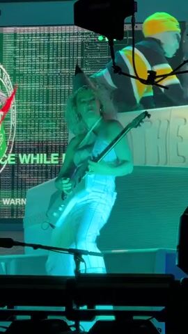 A person wearing a white outfit and a cone-shaped hat is playing a white electric guitar on a stage. The person is positioned in front of a large screen displaying green text and graphics. The stage is lit with blue and green lights, creating a vibrant atmosphere. The guitar has a unique, angular design. The screen behind the person appears to show code or a digital interface, with some text visible such as "ATIONAL TECHNOLOGY" and "LEARNS IS NO AGE".