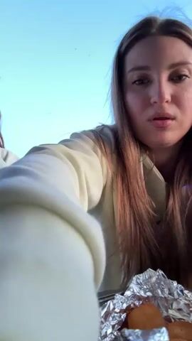 Two young women wearing matching white sweat suits play a hand game next to a tray of food wrapped in foil. They stand in front of a blue sky and a body of water. The woman on the left has curly brown hair and a microphone. The woman on the right has straight brown hair. They repeatedly clap their hands and try to slap each other's hands. The background includes green grass, a body of water, and a few distant structures.