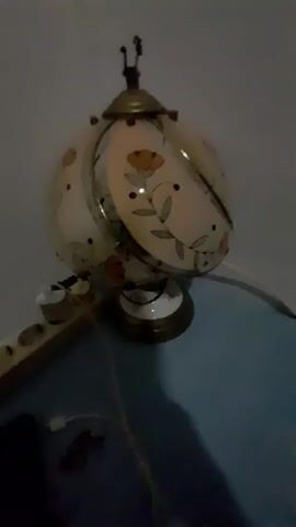A stationary, medium-shot video shows a vintage table lamp with a spherical, cream-colored shade decorated with an orange floral vine pattern. The lamp has a brass-colored base and a decorative finial at the top. It is positioned on a blue surface, possibly a table or shelf, against a pale gray wall. To the left of the lamp, a white electrical outlet strip with several sockets is visible, with white power cords plugged into two of the sockets. One of the cords appears to be connected to the lamp. The lighting is dim, and there is a noticeable shadow cast by the lamp and other unseen objects onto the blue surface. The overall scene suggests an indoor setting, likely a room in a home or office.