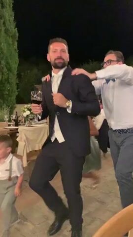 In this video, a group of men are dancing outdoors at night. The man in the foreground is wearing a dark suit and holding a glass of red wine. Another man in a white shirt is behind him, with his hands on his shoulders. A third man in a pink shirt is visible on the right side of the frame. There are trees and string lights in the background, suggesting a festive outdoor event like a wedding reception or party.