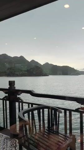 A camera pans across a balcony overlooking a bay dotted with limestone karsts. In the foreground, a wooden chair and a round table are visible. The chair is wet, suggesting it has recently rained. The water is calm, and the sky is overcast. A few boats can be seen in the distance. The overall atmosphere is serene and peaceful.