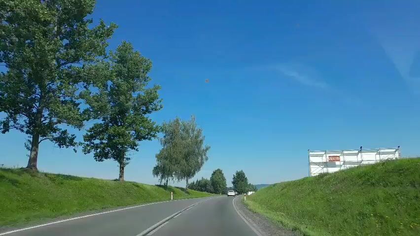 The video shows a view from a car driving on a paved road with green grassy shoulders. There are trees on either side of the road, and in the distance, there are more trees and hills. The sky is blue with a few white clouds. On the right side of the road, there is a structure with scaffolding around it. The car appears to be moving at a moderate speed.