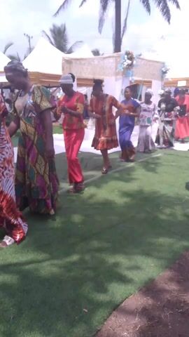 The video shows a group of women dancing in a line on artificial turf. The women are dressed in colorful, patterned outfits. There is a decorated archway behind them, and several white tents are visible in the background. A palm tree stands to the right. The video pans to the side, showing a man taking photos and another man standing next to sound equipment under a tent. The scene appears to be at an outdoor event, possibly a wedding or celebration.