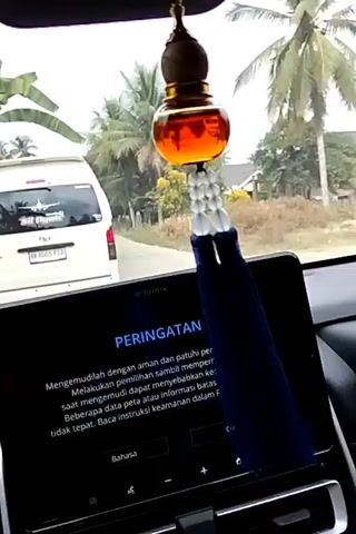 The video captures a view from inside a car, looking forward through the windshield. A decorative car air freshener, featuring a glass bottle with amber-colored liquid and a beaded string with a blue tassel, hangs from the rearview mirror, partially obscuring the view.

Outside the car, a white van with the text "SR Djoyo" and an airplane graphic is visible ahead on a road. The license plate reads "BK 8005 PTD". The setting appears to be a rural or semi-rural area with palm trees and other vegetation. The sky is overcast, suggesting a cloudy day. The road is paved, and there are some buildings visible in the distance.

The car's touchscreen display is also in the foreground, showing a warning message in Indonesian. The text reads: "PERINGATAN" (WARNING). Below this, it says: "Mengemudilah dengan aman dan patuhi peraturan lalu lintas. Melakukan pemilihan sambil memperhatikan layar saat mengemudi dapat menyebabkan kecelakaan serius. Beberapa data peta atau informasi bisa tidak tepat. Baca instruksi keamanan dalam Panduan Pengguna." This translates to: "Drive safely and obey traffic regulations. Making selections while paying attention to the screen while driving can cause serious accidents. Some map data or information may be inaccurate. Read the safety instructions in the User Manual." At the bottom of the screen, "Bahasa" (Language) is visible, along with an "Oke" (OK) button.

The overall impression is that of a driver in Indonesia, possibly on a journey, with a car display providing safety information and a decorative element adding a personal touch. The time of day seems to be daytime, given the natural light.