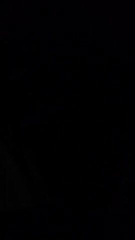 A medium shot shows a person's face illuminated by a soft light source off-camera. The person is wearing a dark shirt and a baseball cap. Their brow is furrowed, and they appear to be speaking or reacting to something unseen. The background is dark and out of focus, with a hint of a wall or structure to the left. The video is low-light and grainy.