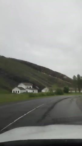 The video shows a view from inside a car driving on a wet asphalt road. The weather is overcast and appears to be raining, as evidenced by the reflections on the road and water droplets visible on the windshield in the foreground. On the left side of the road, there are several white buildings, possibly houses or farm structures, nestled at the base of a steep, green-covered hillside. The hillside has a rocky, reddish-brown cliff face visible near the top. On the right side of the road, more white buildings can be seen, along with a few tall trees. The road curves to the right as the car continues to move forward. A streetlamp is visible on the right side of the road in the distance. The overall impression is a rural landscape under inclement weather conditions.