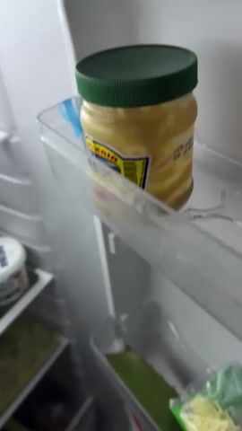 The video shows the inside of a refrigerator. A jar of yellow condiment with a green lid sits on a clear plastic shelf. Below the shelf, more items are visible, including what appears to be grated cheese in a green bag, some ginger roots, and two small white containers. The lighting suggests the refrigerator door has just been opened.