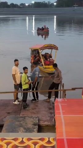 In the foreground, several young men stand on a wooden platform near a body of water. One man in a plaid shirt is holding up a phone to take a picture or video. Another man in a light green shirt is looking down at something in his hands. A man in a blue t-shirt stands next to him. Behind them, a boat with a colorful canopy is docked, carrying passengers. In the background, a smaller red boat with people in it is visible on the water. The scene appears to be an outdoor recreational area, possibly a lake or riverfront, with calm water and a subdued lighting, suggesting it is either dawn or dusk.