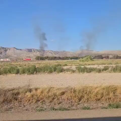 The video shows a landscape with dry, brown vegetation in the foreground and midground. In the background, there are mountains and a clear blue sky. A line of trees runs horizontally across the midground, and several red and white trucks are visible among them. Dark smoke plumes rise into the sky from behind the trees, indicating a fire. The camera appears to be moving slightly, possibly from a vehicle. The overall impression is of a rural or semi-arid environment with an ongoing fire event.