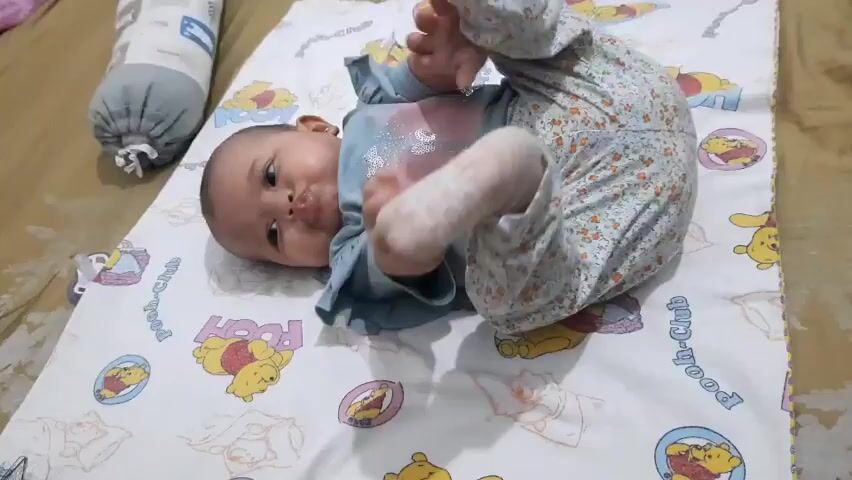 A baby is lying on its back on a Winnie the Pooh themed mat. The baby is wearing a blue shirt with glitter on it and floral patterned pants. The baby is moving its legs and arms, and appears to be playing. There is a grey cylindrical pillow to the left of the baby. The baby is in focus.