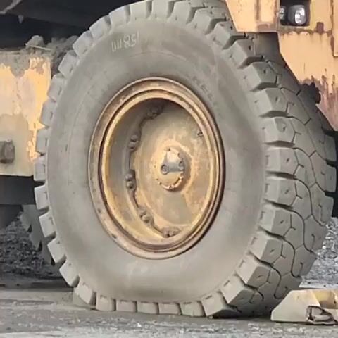 A close-up shot shows the massive tire of a yellow Caterpillar mining truck. The tire is very large, with a thick tread pattern. A partially obscured "CAT" logo is visible on the truck's body. The camera then slowly pulls back, revealing the entire truck, which is also yellow and appears to be in a quarry or mining site. The truck has the number "158" clearly displayed on its rear. The ground is covered in dark gravel. The truck is stationary.