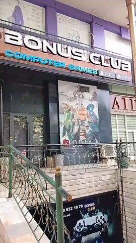 The video shows the exterior of a building with a sign reading "BONUS CLUB COMPUTER GAMES". Below the sign is a large poster for the game Dota 2. A staircase leads down to an entrance with a sign featuring a video game controller and the text "BONUS CLUB". The building appears to be a commercial establishment.