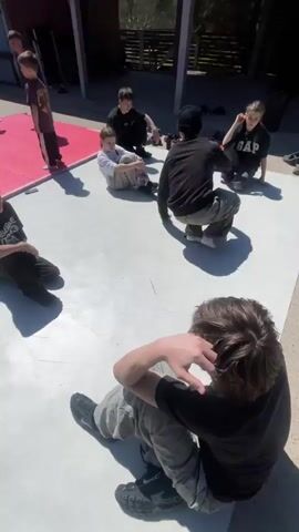 Several people are gathered on an outdoor patio on a sunny day. A group of children and teenagers are seated on a white surface, while an adult in dark clothing seems to be instructing them. Another adult is standing nearby. The patio is partially covered by a roof structure. A red mat is visible near the white surface. The surrounding environment includes trees and a wooden fence. The individuals appear to be engaged in some sort of lesson or training session.