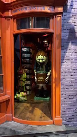 A medium shot captures the entryway of "Weasleys' Wizard Wheezes" in the Harry Potter theme park. The orange-framed doorway reveals a display featuring a figure of a person with an exaggerated smile, holding a bucket labeled "U-No-Puke Sweets". Behind the figure, shelves display products like "Decoy Detonators" and "Hat". A brick wall is visible to the right, while part of the building's facade is seen to the left. The camera remains stationary. The audio captures background chatter.