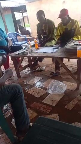 The video shows a group of four men sitting around a wooden table outdoors, possibly under a covered area. The ground is covered with stone tiles and appears wet. On the table, there are several bottles of drinks, some papers, and what appears to be money. Two of the men are seated at the table, one in the center wearing a green shirt and the other on the right in a yellow jacket and a red cap. The other two men are seated on chairs to the left and in the foreground, facing away from the camera. One of these men is wearing a blue t-shirt and shorts, while the other in the foreground is wearing a yellow t-shirt and dark pants. The men appear to be engaged in a discussion or meeting, with some looking at the table and others looking at each other or off-camera.