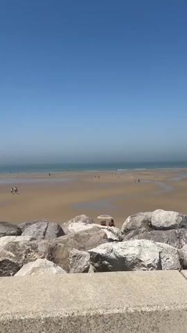 This is a vertical video of a beach on a sunny day. The foreground features large, grey rocks, with a concrete path behind them. Beyond the rocks, a wide sandy beach stretches out to the ocean. The ocean water is a hazy blue-green, with the horizon visible in the distance under a clear blue sky.

Scattered across the beach are several small groups of people. In the mid-ground, two people are sitting in red beach chairs under a colorful umbrella, facing the ocean. Further out on the sand, other individuals and small groups are visible, some walking, some sitting or lying down. A few people are also in the shallow water at the edge of the sea.

The overall scene suggests a relaxed and pleasant day at the beach, likely during the daytime, given the bright sunlight and clear sky. The hazy quality of the water and sky might indicate a warm, possibly humid, day. There is no visible text in the video. The location is identified as Wissant, France.