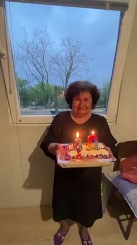 An elderly woman, with short dark curly hair, stands indoors holding a birthday cake with lit candles shaped like "6" and "0." She is wearing a black dress and purple patterned slippers. She smiles at the camera before leaning forward and blowing out the candles. The cake is on a clear tray and has white and red frosting. Behind her is a large window revealing an outdoor scene at dusk or evening, with a cloudy sky and bare trees. The setting appears to be a simple room, possibly a home, in Kosten, Bulgaria.