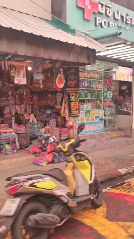 The video shows a street scene with several people. On the left, a pharmacy with an "OPEN" sign is visible. To the right of the pharmacy, there is an ice cream stand with people ordering. Several individuals, including two women and a man, are walking past the ice cream stand and towards the right of the frame. In the background, there are shops and decorations, including red lanterns. The overall environment appears to be a busy street, possibly in a tourist area, with a mix of shops and food vendors.