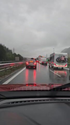 The video shows a view from inside a car on a highway during a rainstorm. The windshield wipers are in motion, smearing water across the glass. Traffic is backed up, with numerous cars and a recreational vehicle visible ahead. Brake lights are illuminated on many vehicles, indicating slow movement or a standstill. The road is wet, reflecting the red brake lights. The sky is overcast and grey, suggesting gloomy weather conditions. The overall scene depicts a typical traffic jam in inclement weather.