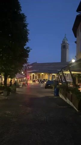The video shows a nighttime view of a European cobblestone street. On the left side, there are trees, potted plants, and outdoor seating for what appears to be a restaurant or cafe, with umbrellas providing shade. A few tables and chairs are visible. Further down the street, there are more outdoor seating areas with lights. In the center of the street, a few cars are parked. In the background, a large building with a tall bell tower is illuminated. On the right side of the street, there is a building with outdoor dining tables and umbrellas, with a few people seated and dining. Bicycles are parked in front of this establishment. The sky is a deep blue, indicating it is dusk or early night. The overall atmosphere is that of a quiet evening in a town square with dining establishments open.