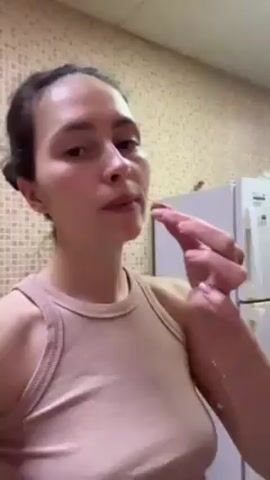 A woman stands in a kitchen, holding a small, popcorn-shaped piece of food between her thumb and index finger. She brings it to her mouth and eats it. She is wearing a light pink tank top. A white refrigerator is visible behind her. The walls are tiled. Her dark hair is pulled back.