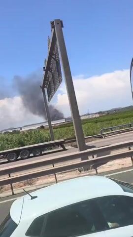 This video shows a fire sending a large plume of black smoke into the air. The fire appears to be in an industrial area, with several large buildings visible in the background. A highway with several cars and trucks is in the foreground, with traffic moving slowly. The sky is blue and there are some clouds. The video is taken from a moving vehicle.