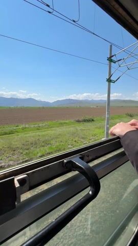 The view from a train window shows passing fields and distant mountains under a bright blue sky with scattered clouds. The foreground is dominated by the train's window frame, a black handle, and glimpses of the trackside gravel. A hand rests on the window frame as the train moves, the landscape a blur of green grass, brown plowed fields, and occasional structures such as utility poles.