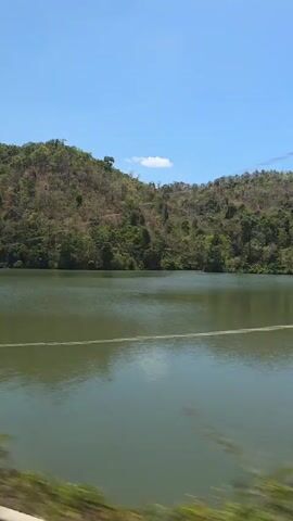 A daytime video shows a serene natural landscape featuring a large body of water, likely a lake or reservoir, bordered by rolling hills. The camera appears to be moving, possibly from a vehicle, offering a panoramic view.

The water, a murky green-brown color, occupies the foreground and midground, reflecting the clear blue sky. A thin, light-colored line or rope stretches across the water horizontally in the distance. Along the near bank, lush green vegetation can be seen, contrasting with the water.

The background is dominated by a series of hills and mountains covered in a mix of verdant green trees and sparser, drier-looking foliage, suggesting a season or ecological characteristic. A clear, bright blue sky with a single small white cloud stretches overhead, indicating sunny weather.

The video is accompanied by an abrupt, metallic clanking sound followed by a low, consistent mechanical hum, suggesting nearby machinery or the movement of the recording device. There is no visible text in the frame. The scene evokes a tranquil, rural setting, consistent with the provided context of Phan Sơn, Vietnam.