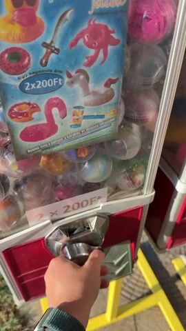 A hand is shown interacting with a gumball machine. The machine has a red base and a clear plastic top filled with colorful gumballs inside clear plastic capsules. A sign on the machine displays images of inflatable toys like a duck, a sword, an octopus, a unicorn, a flamingo, and a life preserver. Text on the sign indicates the price is "2x200Ft." The hand inserts coins into the machine's coin slot, then turns a knob. A gumball capsule is dispensed from the bottom of the machine, and the hand reaches to retrieve it. The machine is outdoors, with sunlight casting shadows and illuminating the scene. A yellow stand supports the machine.