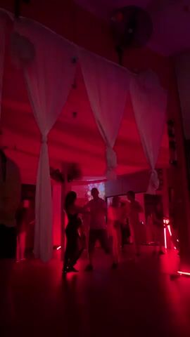 The scene is an indoor dance party under red lighting, with white drapes hanging from the ceiling and a fan visible in the upper-right corner. Several people are dancing, illuminated by the red light, with a couple dancing in front of a mirrored wall. Additional decorations are placed around the room, contributing to the party atmosphere.