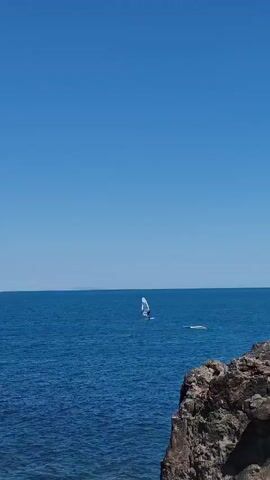A wide shot shows a vast expanse of calm, deep blue ocean under a clear, bright blue sky. In the middle distance, a single windsurfer is visible, their white sail catching the light. To the right of the windsurfer, a person is swimming or treading water, with their head and shoulders visible above the surface. In the foreground, a rugged, rocky cliff face juts out into the water on the right side of the frame, with some dark green vegetation growing on its upper edges. The overall scene depicts a serene day at sea with water sports activities.