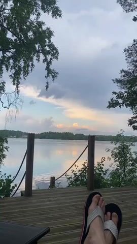 A first-person perspective video shows a person relaxing on a wooden deck overlooking a calm lake. The person is wearing flip-flops. In the background, trees line the shore and the sky is filled with soft, colorful clouds. To the right of the frame, a wooden hot tub with a metal chimney is situated on the deck. A paddle is leaning against the hot tub. The video pans slightly to the right, revealing more of the trees and the hot tub.