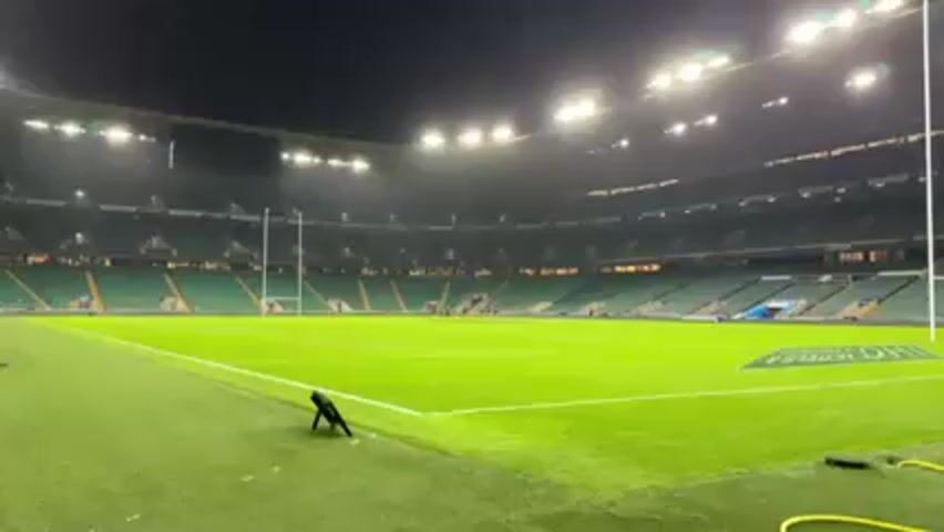 The video shows a wide shot of a large, mostly empty rugby stadium at night. The field is brightly lit and green, with white lines marking the playing area. There are two tall rugby goalposts at either end of the field. The stadium seating is green and mostly unoccupied. Above the seating is a large screen and numerous stadium lights. In the foreground, there is some equipment on the edge of the field, including a black camera tripod, a yellow hose, and some black cases. Two men briefly appear in the frame, standing near the equipment before walking out of view. The camera is static throughout the video.