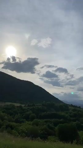 The video shows a hillside covered in green trees, with the sun shining through a cloudy sky. In the background, snow-capped mountains are visible. The camera pans slowly from left to right. The weather appears to be partly cloudy. There are no people visible.