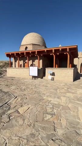 The video shows a traditional brick building with a domed roof and a wooden portico. The building is set in a courtyard paved with irregular stone tiles. A clear blue sky is visible above. Two informational signs stand in front of the building, one larger than the other. The larger sign has text and images, while the smaller one has a plaque. The building appears to be an ancient structure, possibly a historical monument or a religious site. The environment is arid, with dry vegetation and sandy soil visible in the background. The lighting suggests it is daytime, possibly late morning or early afternoon. There are no people visible in the video.