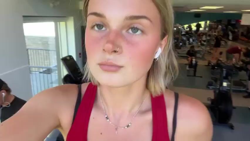 A young woman with blonde hair and light eyes is shown exercising in an indoor gym. She is wearing a red tank top with black straps, black shorts, and light-colored athletic shoes. White earbuds are in her ears, and she wears a silver necklace.

The video features close-up shots of her face, which appears slightly flushed, indicating physical exertion. She maintains a neutral, focused expression. An overhead shot shows her from the waist down, actively using a treadmill. Her feet are moving on the treadmill belt. The treadmill's console is visible, displaying some digital readings and green and red indicator lights, though specific numbers or text are not clearly legible. The background in all shots is somewhat blurry, revealing other gym equipment, confirming the setting as a fitness facility. The provided location context is Whistler, Canada.